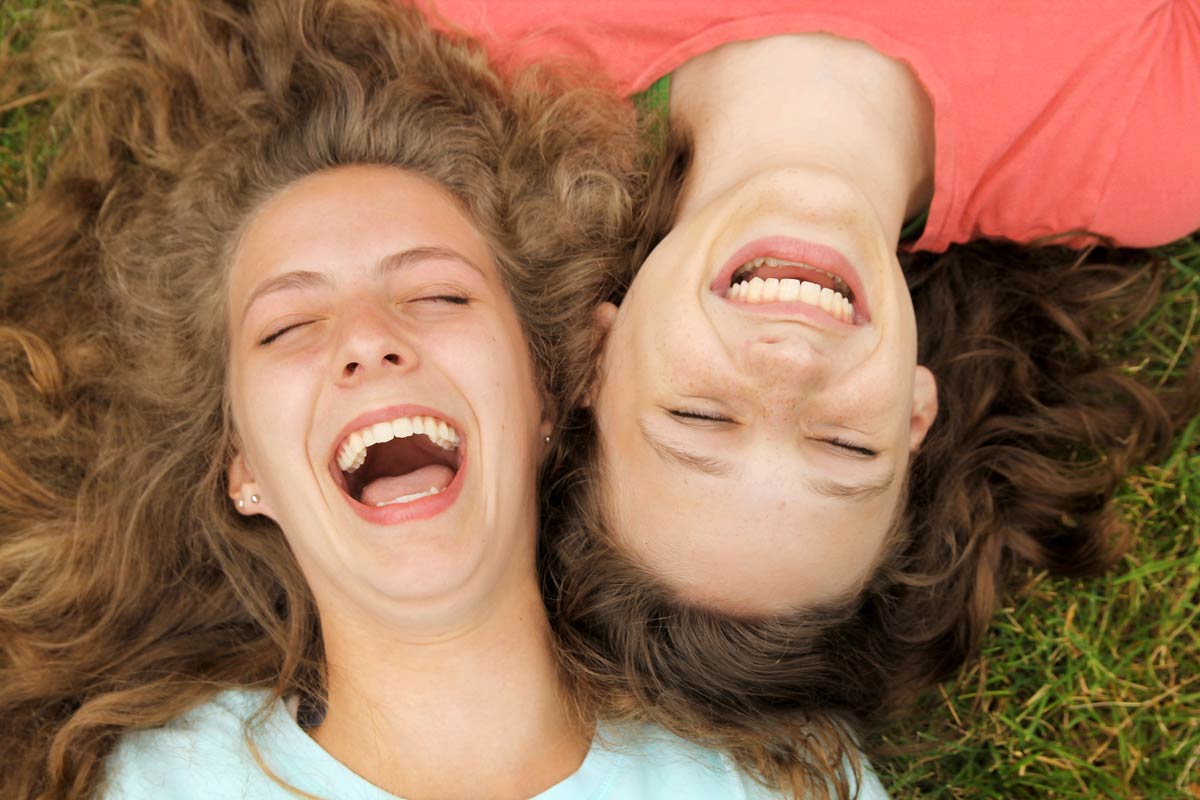 Two young women lying in the grass, laughing joyfully, visually representing the mood and well-being linked to what terpenes make you laugh.